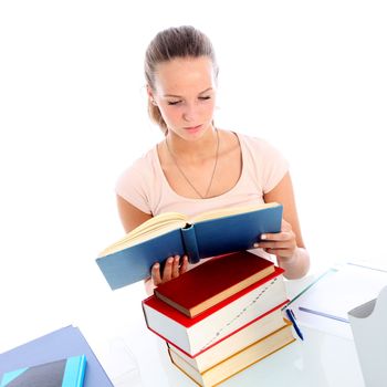 Dedicated young female student sitting at her desk with a pile of books immersed in reading a textbook while studying 