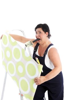 Exasperated woman hanging wallpaper with a brush full of glue paste clenched in her mouth as she holds the roll of paper in both hands Exasperated woman hanging wallpaper with a brush fulll of glue paste clenched in her mouth as she holds the roll of paper in both hands 