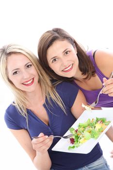 Two pretty women friends standing close together enjoying a healthy salad eating off a shared plate isolated on white Women friends standing close together enjoying a healthy salad eating off a shared plate isolated on white