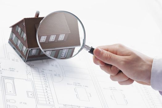 Concept image of a home inspection. A male hand holds a magnifying glass over a miniature house. 