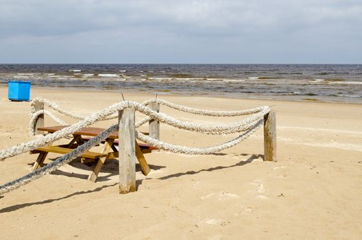 Fence made ??of logs and thick rope on seaside beach sand. Wooden table and benches. Blue trash container.
