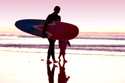 Female surfer and her daughter walking in the beach at the sunset