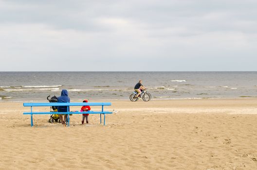 woman with kid sit on bench and bicycle go near sea coast line on beach.