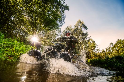 Quad rider through water stream in the forest against sunlight.