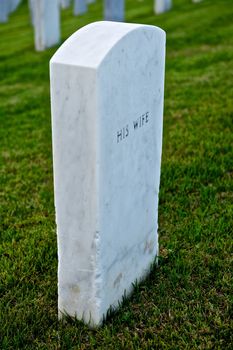 White marble headstone or grave marker with "HIS WIFE" engraved in black lettering in a cemetary