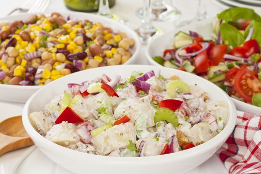 An assortment of salads on a buffet table. Potato salad, bean salad and fresh mixed salad arranged on a white table with glasses, cutlery and plates.