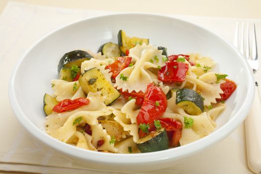 A bowl of pasta bows or farfalle with roast courgettes or zucchini, red pepper and tomato with olive oil and herbs.