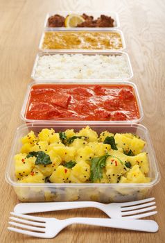 A selection of Indian takeaway food in plastic containers on a wooden table. Aloo saag (potato spinach curry), chicken tikka masala,basmati rice  chicken bhoon or  bhuna, and onion bhaji or pakora.