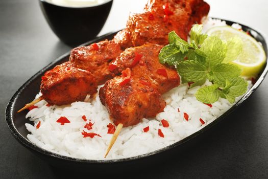 Tandoori chicken kebabs on a bed of rice, garnished with mint and lime, served with yoghurt, on a dark background.