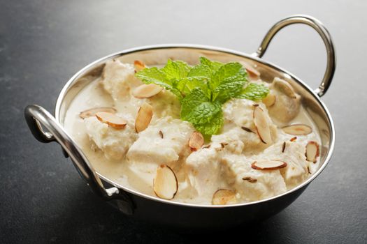 Mild and creamy chicken korma, in a balti dish, garnished with mint, almonds and toasted cumin seeds, on a dark background.