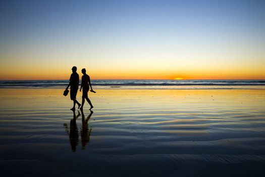 Young couple in silhouette against sunset walking at edge of sea on Cable Beach Broome Western Australia. Both are barefoot and carrying shoes. The end of a perfect day.