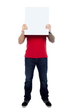 Full length portrait of casual man hiding his face behind blank white ad board