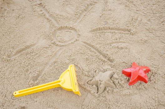 Sun drawn in sand with beach toys next to it. Concept of fun summer holidays.