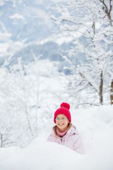 Happy girl in red hat smiling and having fun outdoors on snowing winter day, making snowball and loking at the camera