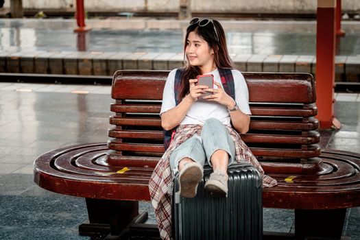 Young hipster woman waiting on the station platform with backpack. Travel concept. asian girl using smartphone at train station.