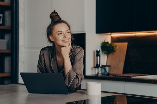Young happy freelancer lady in cozy pajamas working from home and checking emails on her laptop computer in morning while having cup of coffee in stylish kitchen interior
