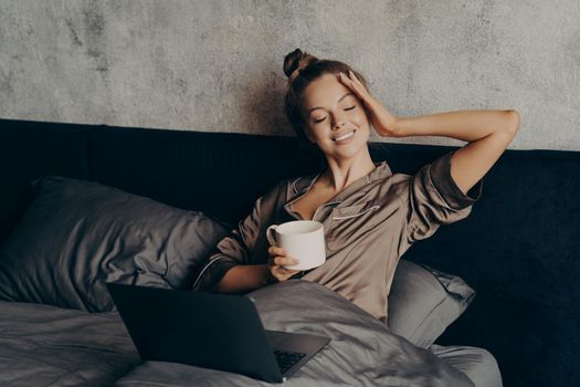 Charming relaxed young woman lying in bed at home with laptop and having morning cup of coffee, watching her favorite TV show online on computer, spending leisure time at home on weekend