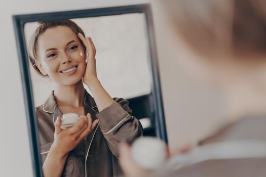 Positive happy young woman in pajama smiling, applying facial cream and looking in mirror, attractive lady doing morning beauty routine in bedroom at home. Skin care concept