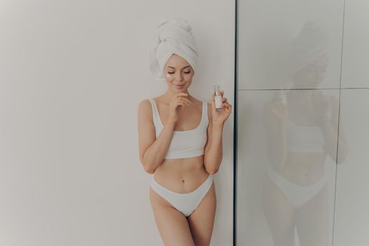 Image of young girl posing with cosmetic moisturizing face cream or serum in hand isolated over light wall in bathroom, wears towel wrapped on head after morning shower. Beauty and skincare concept