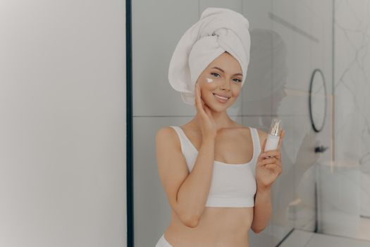 Positive young woman with head wrapped in towel smiling while applying facial cream, attractive lady using cosmetic product while doing beauty morning routine in bathroom at home. Skin care concept