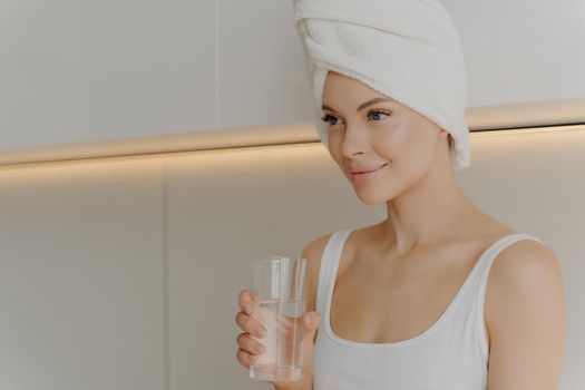 Preventing dehydration. Image of young beautiful woman with fresh and glowing skin drinking water in morning straight after shower with towel wrapped on her head sitting in light colored kitchen