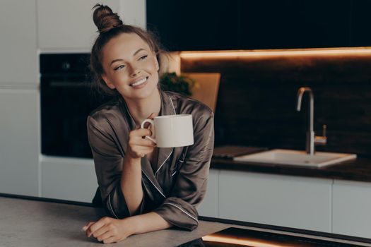 Happy smiling young woman in homewear enjoying aromatic morning coffee in modern kitchen after waking up before busy start of working day, positive female spending leisure time at home