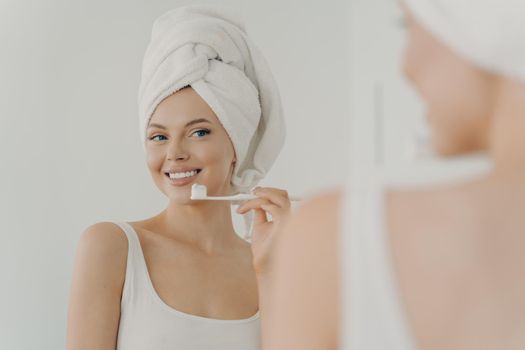 Beautiful young woman with healthy perfect smile brushing teeth and looking in mirror, attractive young female wearing white bath towel on head standing in bathroom at home. Oral hygiene concept