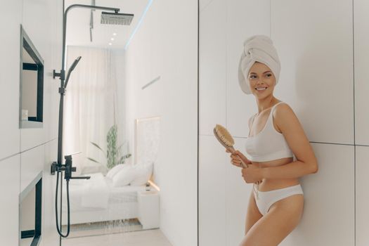 Slender happy female model posing with head wrapped in towel leaning against bathroom wall and holding wooden brush, preparing for morning shower and hygiene routine. Women beauty and body care