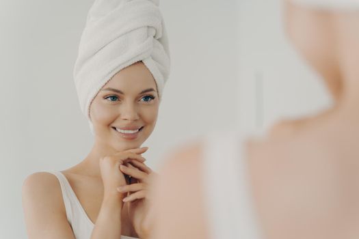 Positive healthy young woman smiling while applying facial cream reflecting in mirror, happy attractive lady putting moisturizing nourishing cream during morning beauty routine in bathroom