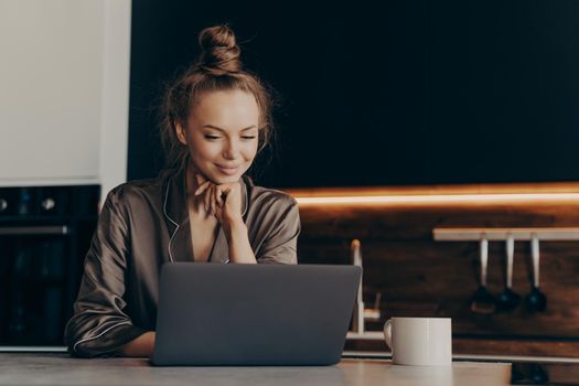 Young female freelance worker in pajama drinks coffee in morning for successful productive working start while sitting in kitchen with laptop and checking emails. Remote work and freelance concept