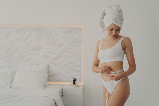 Attractive young girl with sports figure measures her waist with tape centimeter, wears white underwear and bath towel wrapped on head after morning shower routine. Weight loss and dieting concept