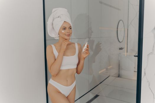 Attractive young adult woman applying facial cream, beautiful healthy lady with head wrapped in towel putting moisturizing lifting nourishing day cream on clean soft skin in bathroom at home