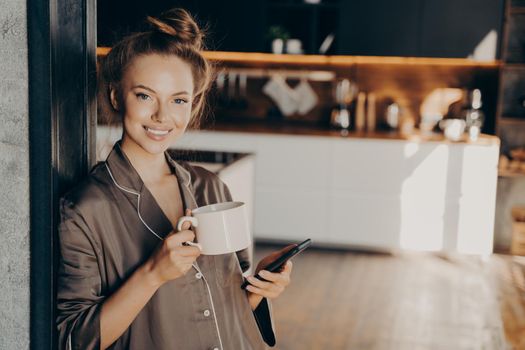 Young positive business woman wears silk brown pajama smiling while holding smartphone in hand and drinking coffee after waking up in morning at home before going to work, standing in bedroom