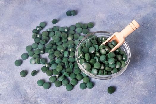Green pills of spirulina or chlorella on a gray-blue plaster background in a glass bowl and in a wooden spoon. Healthy superfood diet and detox nutrition concept.