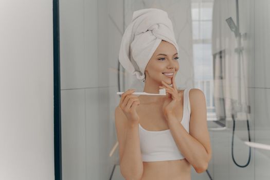 Oral hygiene concept. Healthy beautiful woman with white bath towel on head brushing teeth during morning routine, pretty young female with healthy toothy smile standing in bathroom after shower