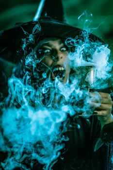 Portrait of witch with awfully face in creepy surroundings and smoky green background drinks magic potion from the goblet.