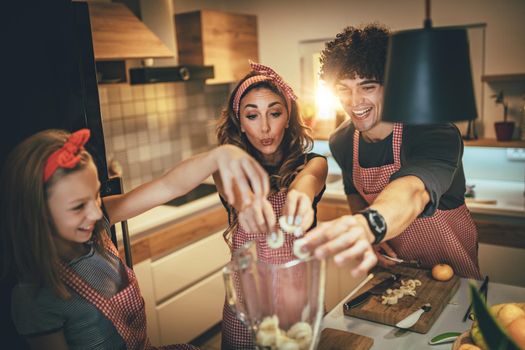 Happy parents and their daughter preparing healthy meal together in the kitchen and putting banana slices in the blender.