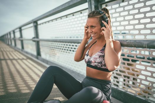 Young fitness woman is resting after hard training on the river bridge and putting her earphones.