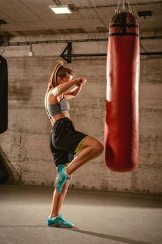 Muscular woman punching with leg a boxing bag on cross fit training at the gym.