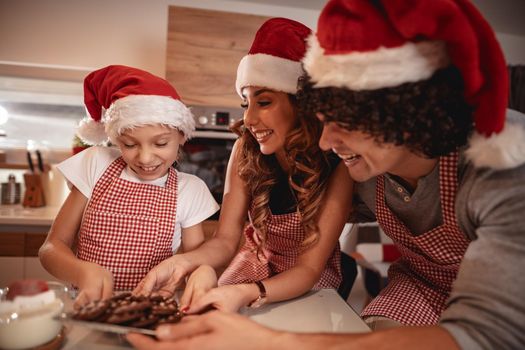 Happy parents and their daughter with santa's cap are preparing meal together in the kitchen. Little girl is offering cookes to her mother and father.