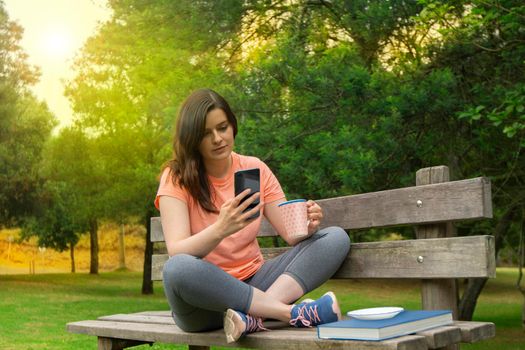 Beautiful young Hispanic woman sitting on a wooden bench in the park dressed in sportswear with a cup of coffee in her hand reading a message on her smartphone with green trees background