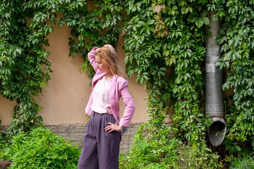 Young elegant beautiful blonde millennial with long hair in pink clothes stands and straightens her hair near the wall of an old building overgrown with wild girlish grapes. Selective focus.