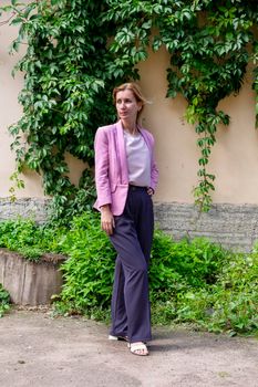 Young elegant blonde millennial with long hair in pink clothes stands and straightens hair near the wall of an old building overgrown with wild girlish grapes. Selective focus. Vertical orientation.