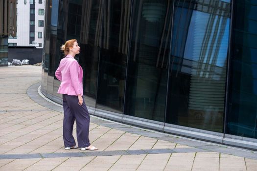 Young blonde woman with tailored hairstyle in pink jacket stands in front of modern office building made of glass and steel and looks at her reflection. Profile view. Selective focus.