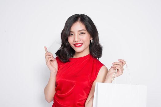 Beautiful asian woman wearing a red dress with shopping bag and holding credit card standing over white.