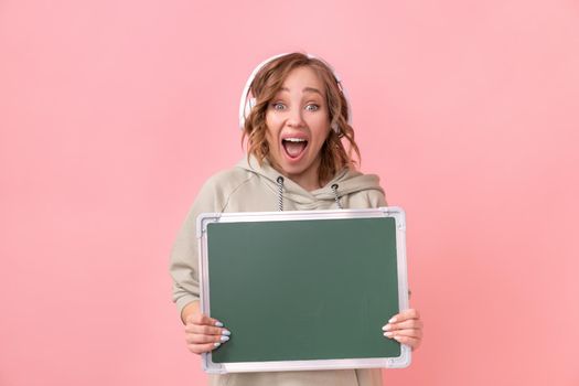 Woman holding empty chalkboard over pink background Happy Caucasian female 30 years old dressed headphones oversize hoodie hold blank green board sign Studio shoot Advertising Promoting Presentation