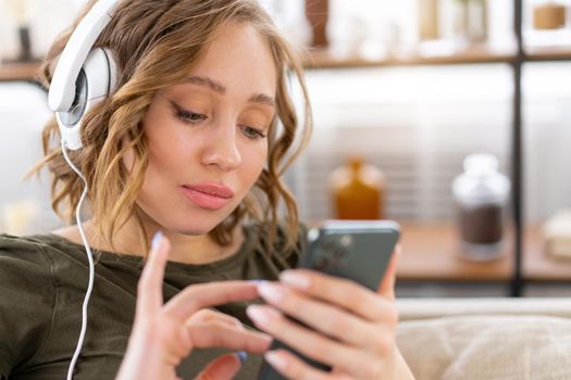 Woman listen music headphones holding smartphone in hand Caucasian female enjoy podcast or audio books while restin on couch close up portrait