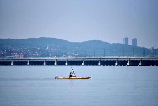 a man floats on a yellow kayak on the sea along a low-water bridge. the city is slightly visible in the background.