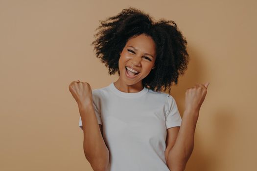 Young euphoric happy african american woman in white tshirt clenching fists and celebrating achievement, overjoyed dark skinned female making yes gesture while posing isolated on yellow background