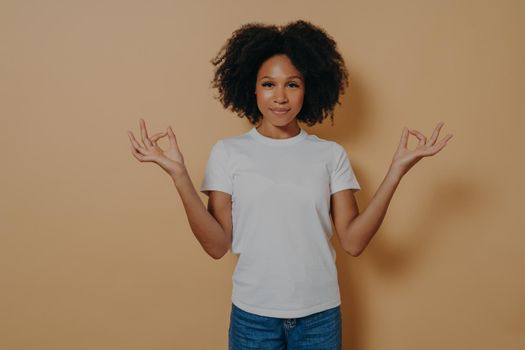 Mindfulness concept. Peaceful afro american young woman in casual outfit keeping hands in mudra gesture and looking at camera with tender smile, being calm and relaxed, isolated on beige wall